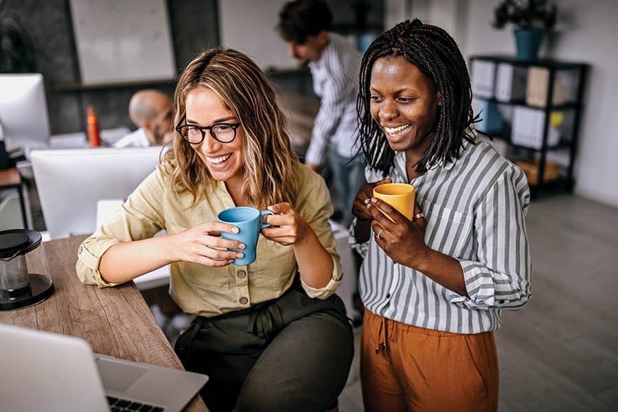 Female colleagues are commenting and enjoying laptop content at the work in a friendly atmosphere in the office while drinking coffee